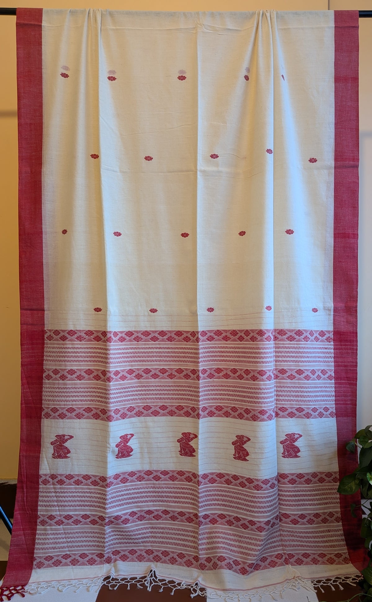 A cream-colored cotton saree or fabric panel with a wide red border, red dotted motifs, and a decorative red patterned border near the bottom, displayed hanging flat.